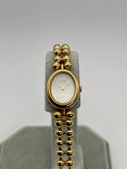 Gold watch with a white face on a gray stand against a light gray background