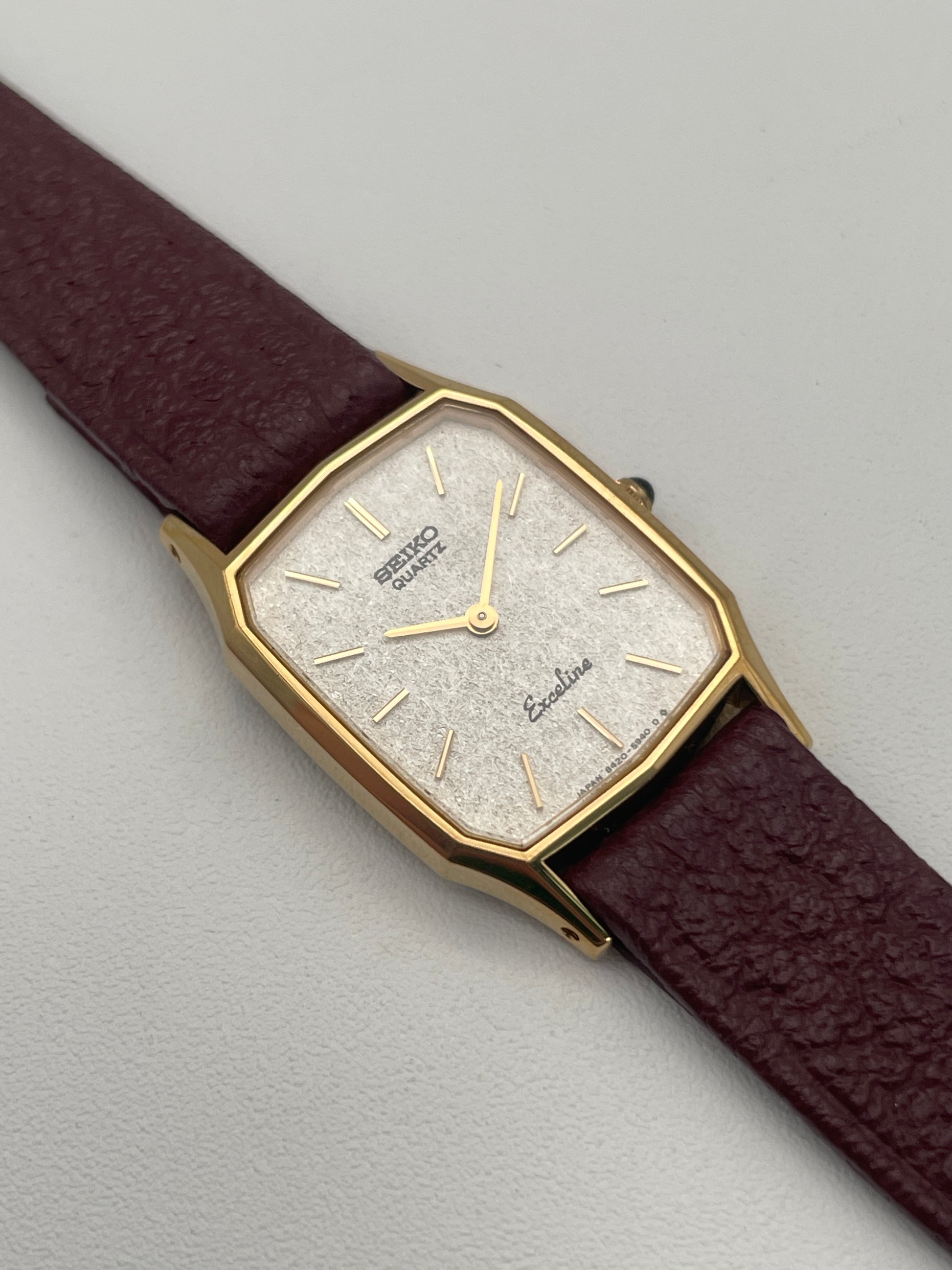 Front flatlay view of the Seiko Exceline Decagon Ladies Watch in gold-tone case with silver textured dial, burgundy leather strap.