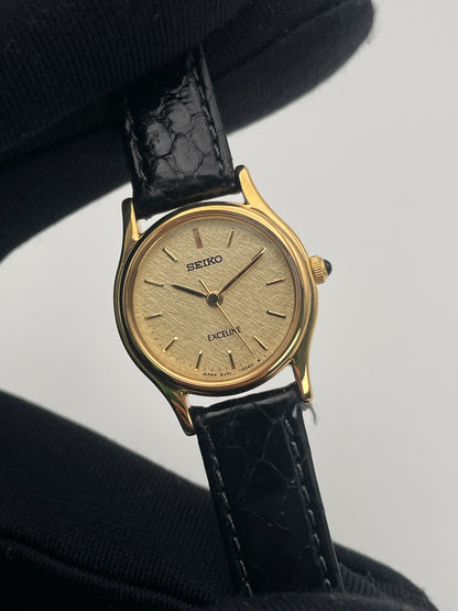 Close-up of Seiko Exceline gold watch held by a gloved hand, showcasing the detailed textured dial.