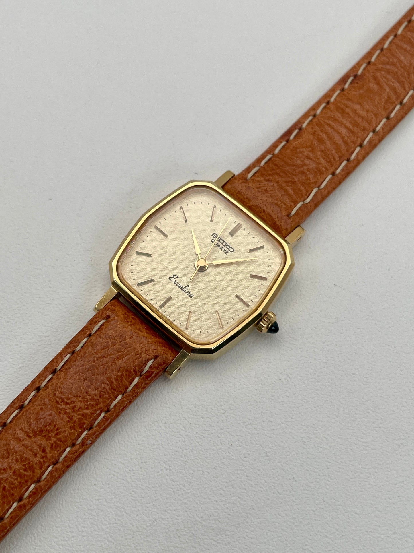 Diagonal view of Seiko Exceline vintage women's watch in gold with tan leather strap, emphasizing the cloth-textured dial and octagonal design.