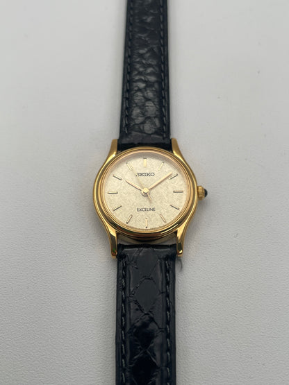 Front view of Seiko Exceline Ladies Watch in gold with a textured dial and black leather strap.