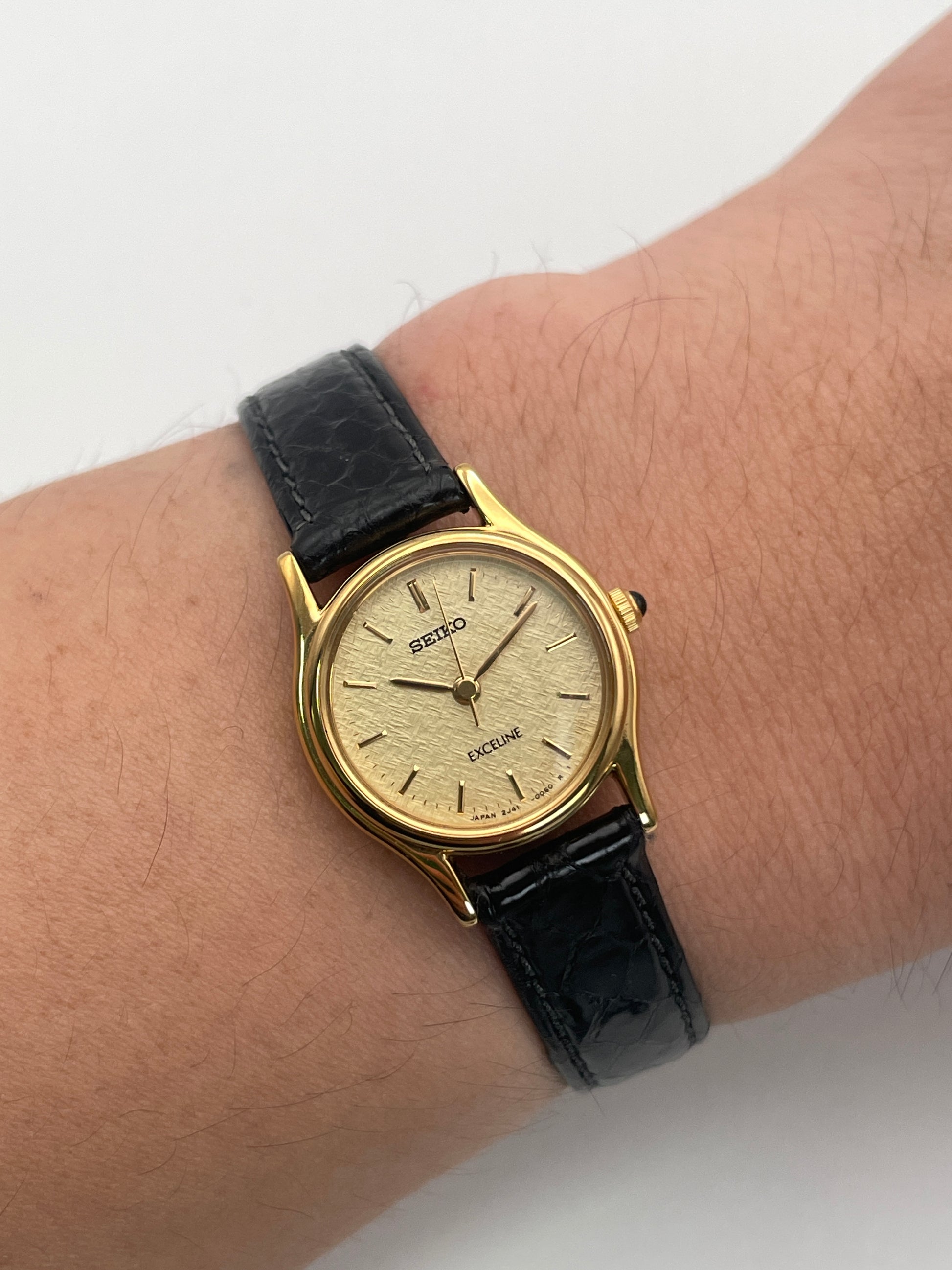 Wrist shot of Seiko Exceline gold watch with a textured dial on a black leather strap.