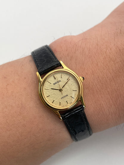 Wrist shot of Seiko Exceline gold watch with a textured dial on a black leather strap.