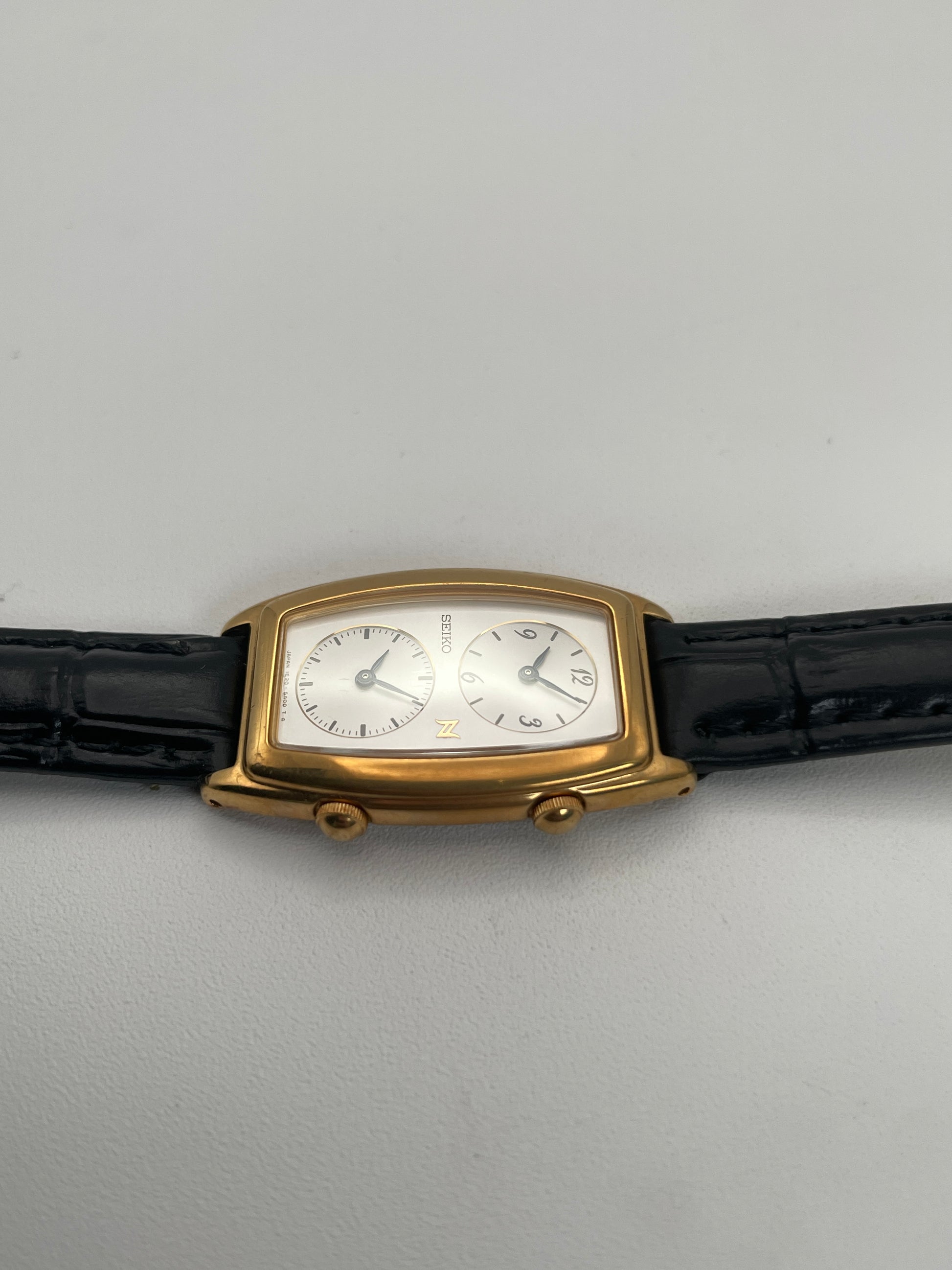 Side crown view of Seiko Noie Dual Time watch with dual crowns on the gold-tone rectangular case and black leather strap.