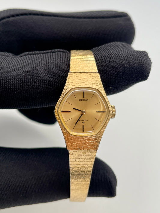 Close-up view of a vintage Seiko ladies' watch with a gold-tone textured case and bracelet, featuring a simple gold dial with thin black hour markers and hands.
