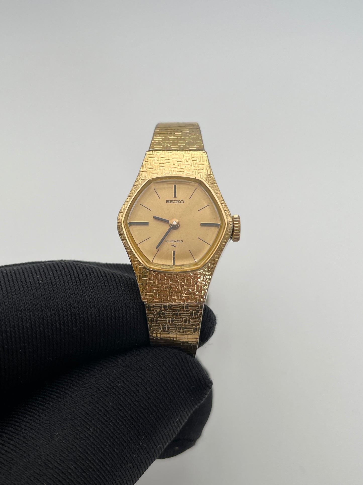 Front view of a vintage Seiko ladies' watch held in a gloved hand, featuring a gold-tone textured case and bracelet with a simple gold dial, black hour markers, and hands.

