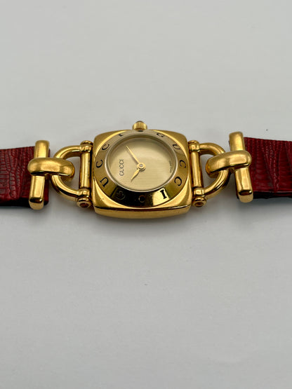 Side profile of a Gucci 6300L ladies' watch, emphasizing the gold-tone case and the connection to the red leather strap.