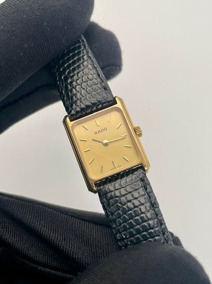 Vintage Rado Tank watch in gold case with a rectangular dial and black leather strap, close-up view in hand.