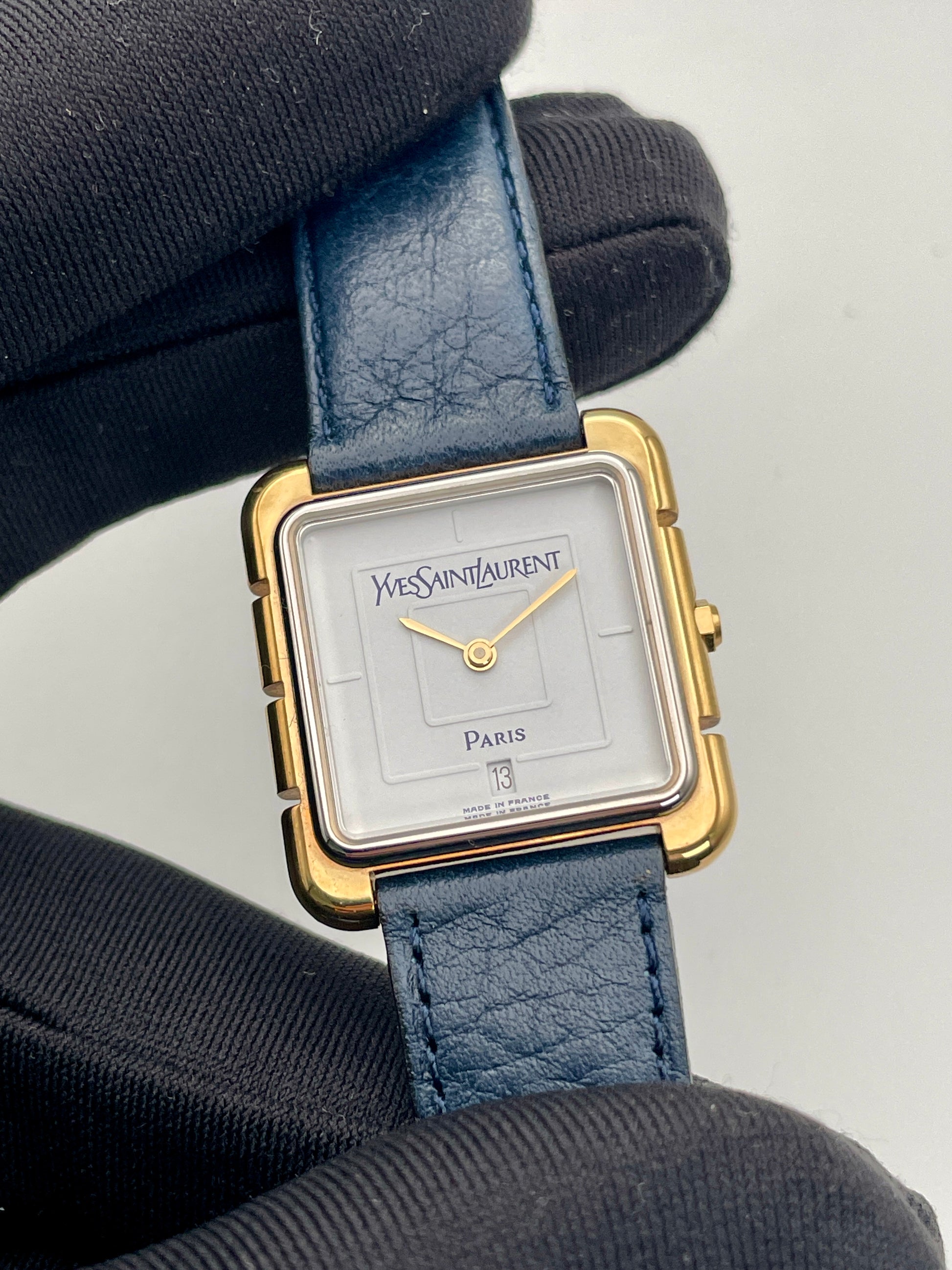 Close-up view of Yves Saint Laurent 3D Square watch's white dial, gold hands, and blue leather strap.