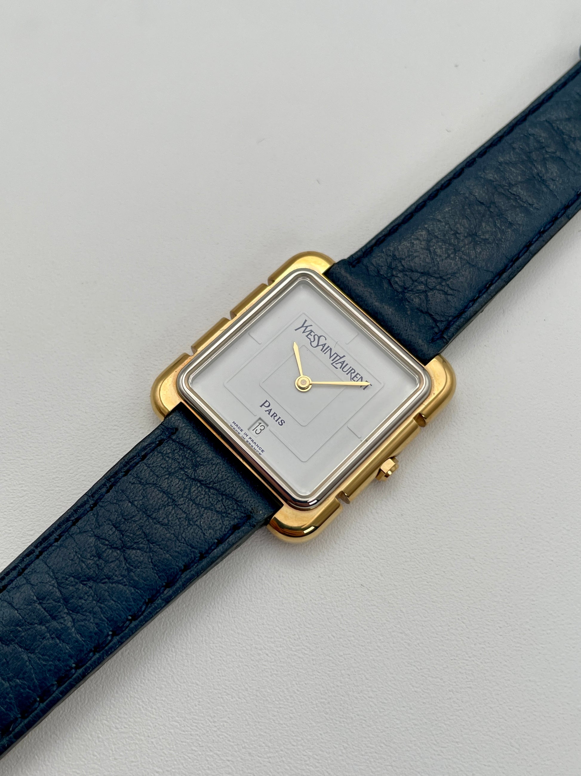 Diagonal view of Yves Saint Laurent 3D Square watch with gold case, white dial, and blue leather strap.