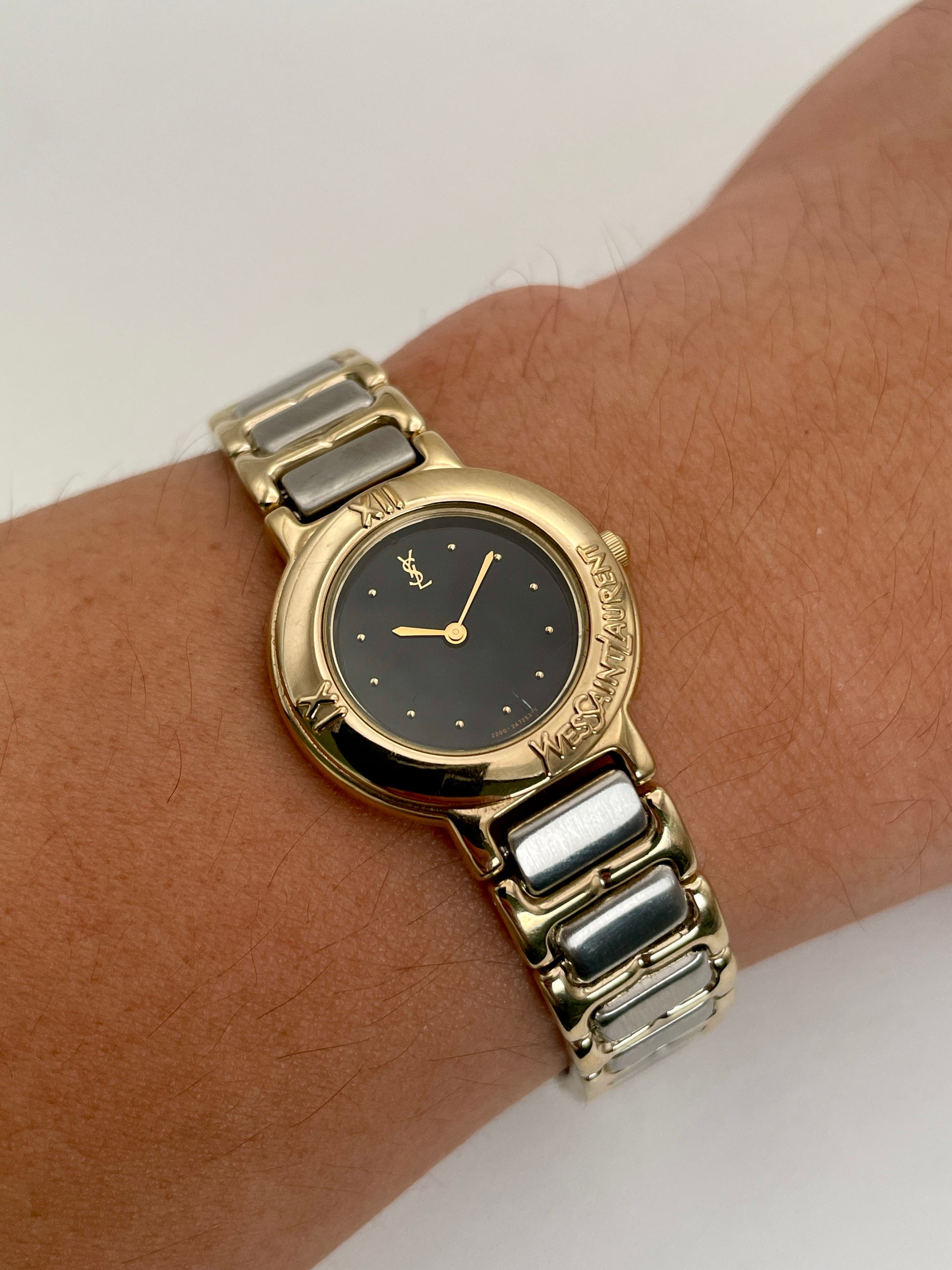 Wrist shot view of Yves Saint Laurent Pour Femme vintage ladies watch with gold case, black dial, and two-tone bracelet.