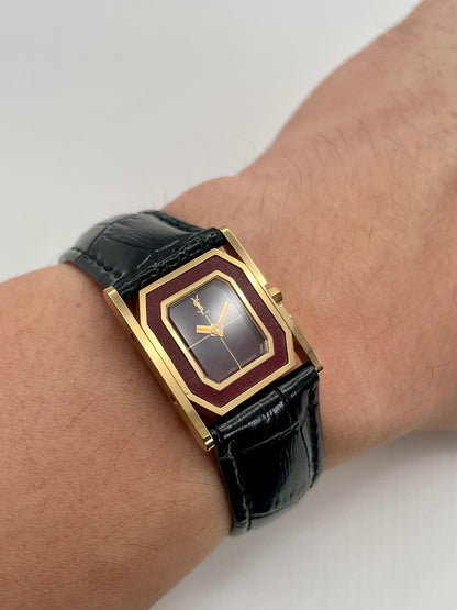 Wrist shot of the Yves Saint Laurent Tank gold watch with burgundy dial and black strap